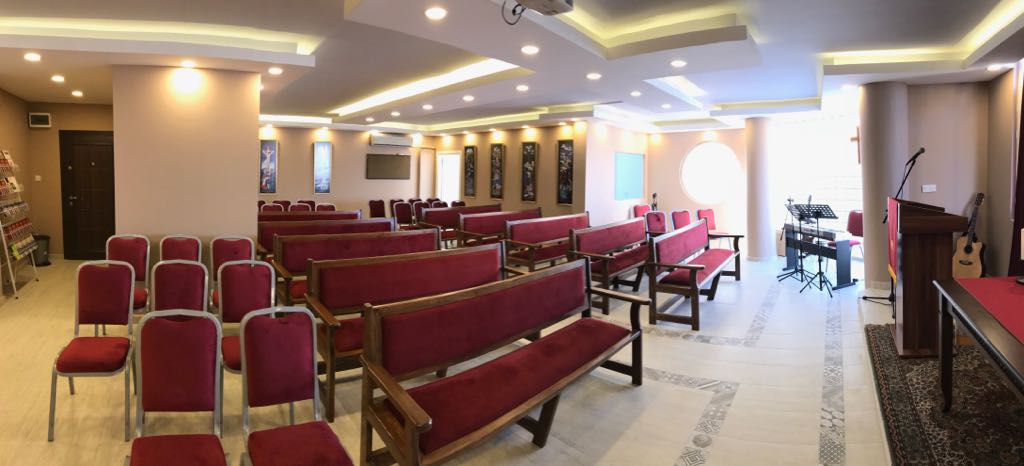 İbadet salonu, arka köşeden: bordo kadife sıralar ve sandalyeler, sağda ahşap kürsü, klavye ve gitar