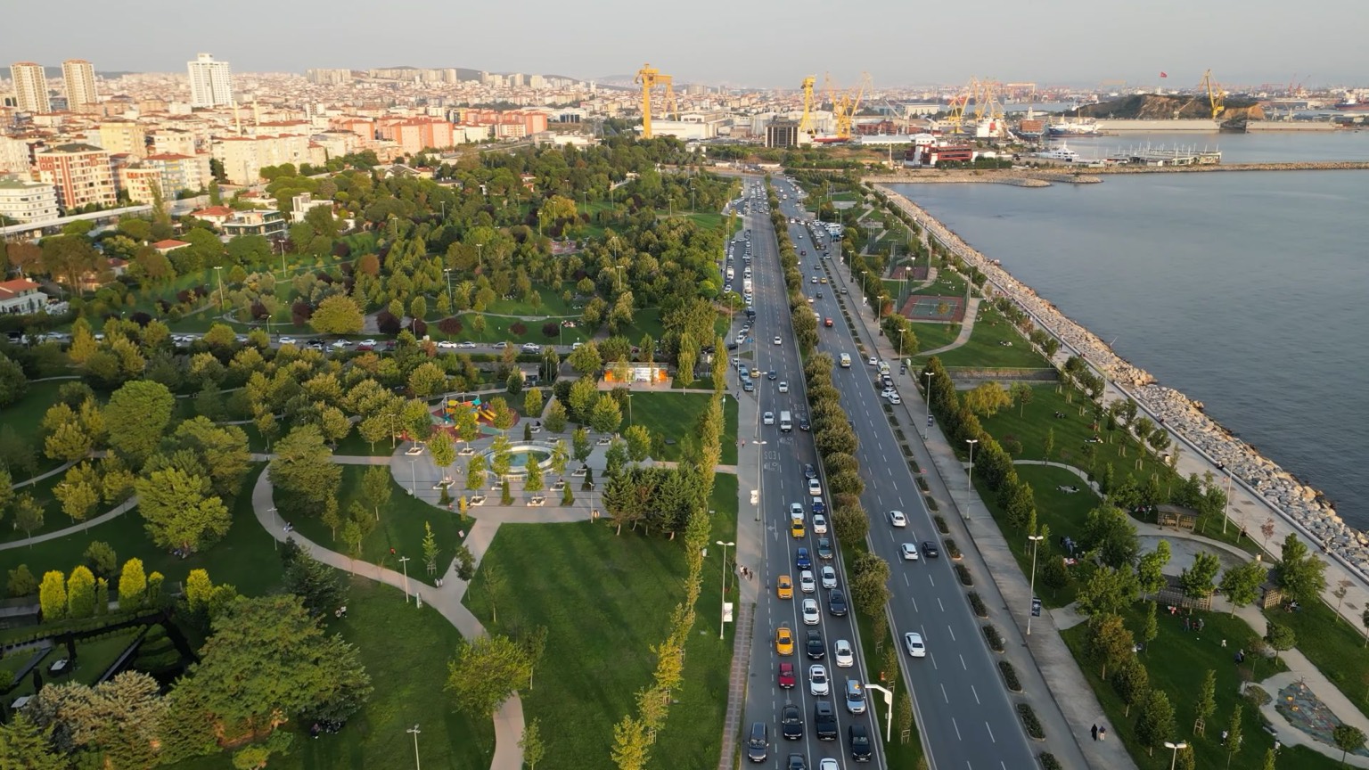 Pendik sahilinde kıyı parkı: ağaçlı yeşil alan ve yürüyüş yolları, yanından geçen sahil yolu, arkada tersane vinçleri ve marina